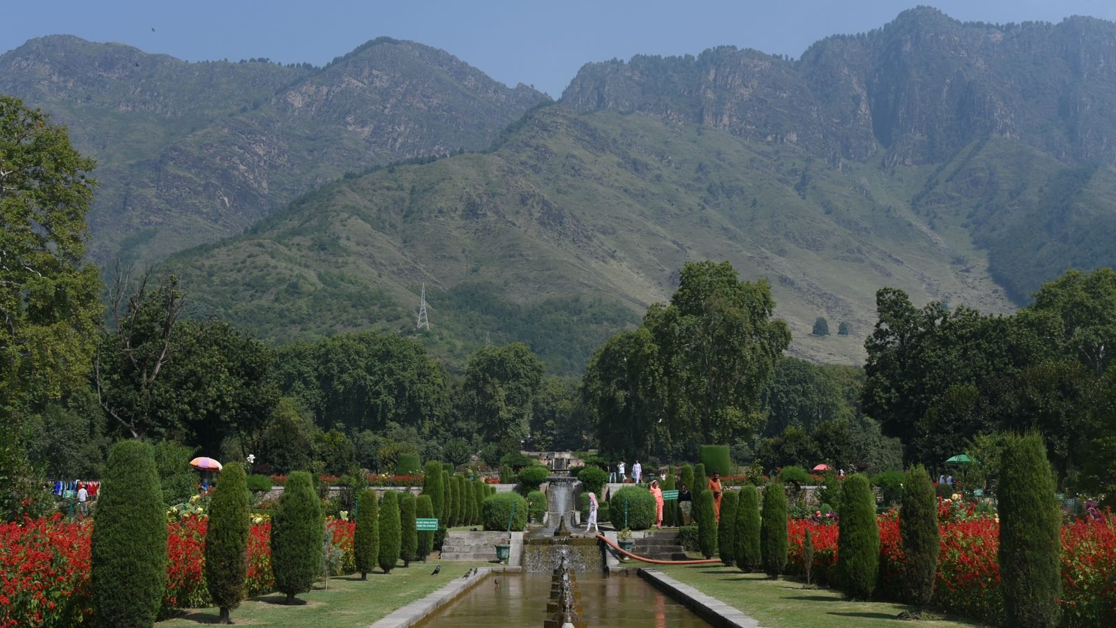 Mughal garden in Srinagar with a fountain channel, red flower beds and clipped cypress trees below green hills