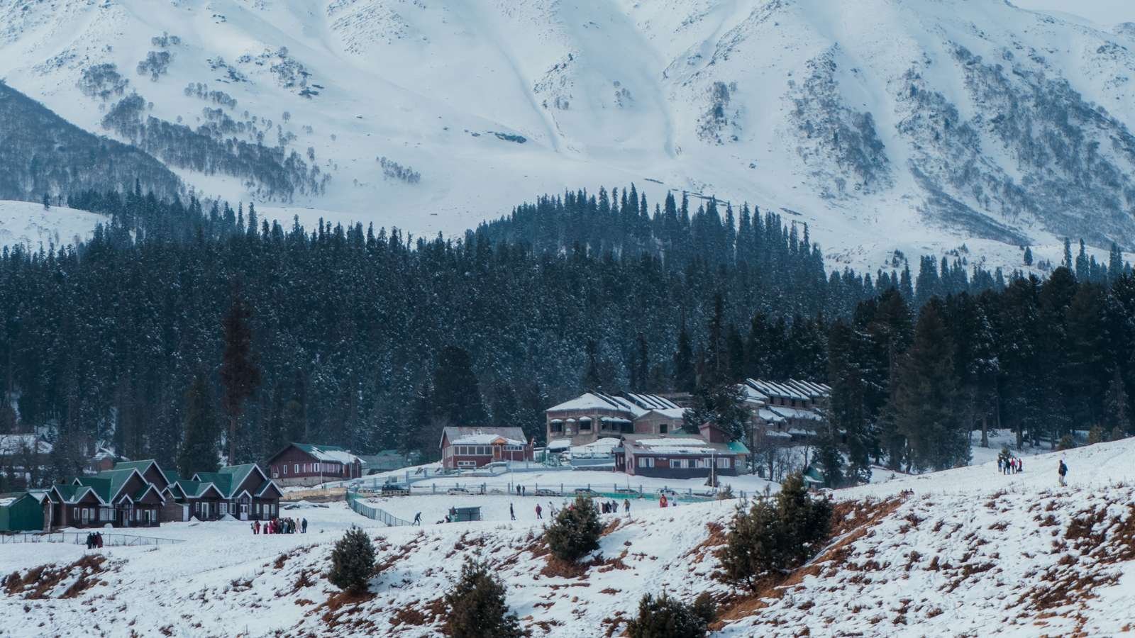Snow-covered Gulmarg meadow with wooden cabins and visitors below pine forest and a steep snowy mountain